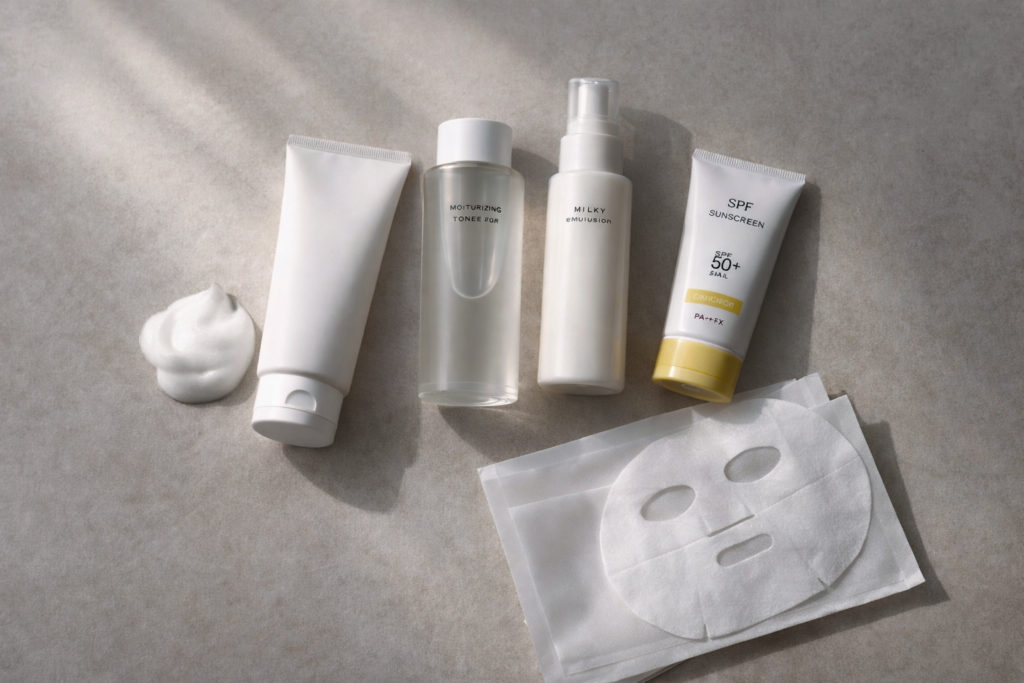 Vista cenital minimalista de una rutina de cuidado facial: limpiador, tónico/loción, emulsión y protector solar SPF 50+ sobre una superficie beige texturizada, junto a varias mascarillas tipo ‘sheet mask’; luz suave y sombras delicadas.